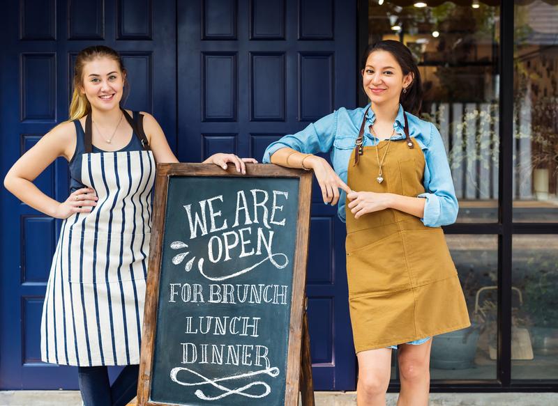 Two women in aprons standing outside a café next to a ‘We are open’ chalkboard