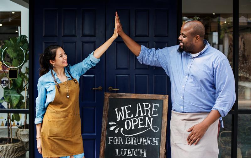 Two café owners in aprons high-fiving beside a ‘We are open’ chalkboard outside their shop