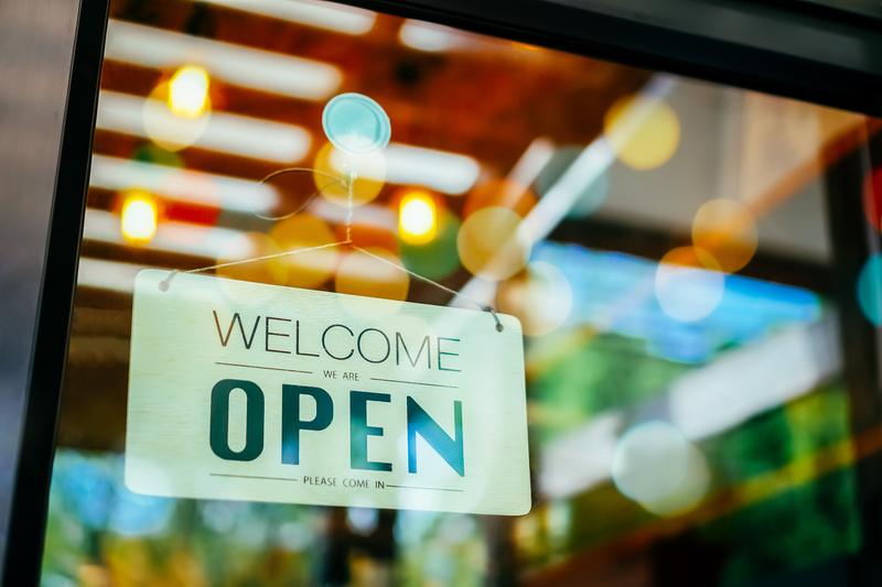 A ‘Welcome, we are open’ sign hanging in a shop window, with soft blurred lights behind